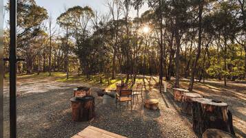 An outdoor seating area with wooden benches and a fire pit, surrounded by trees and gravel.