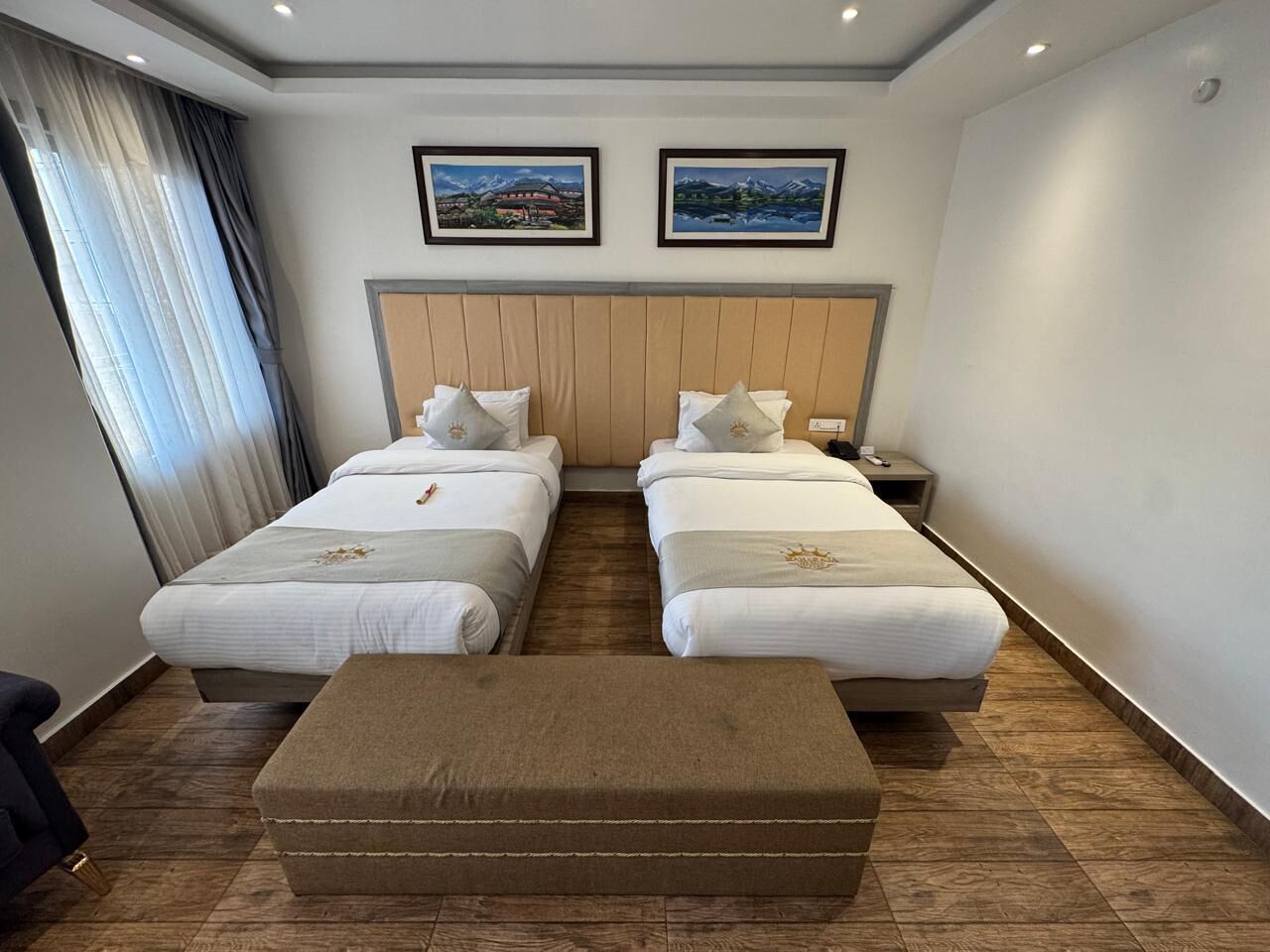 A hotel room with two single beds, a wooden headboard, a large window with curtains, and two framed pictures on the wall.