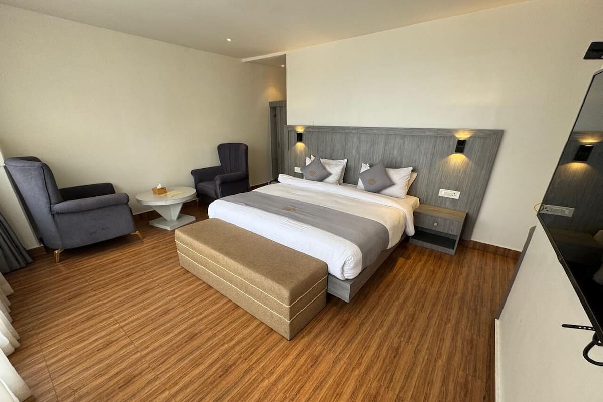 A modern hotel room with a wooden floor, a large bed with a grey headboard, a bedside table, a chair, and a bench.