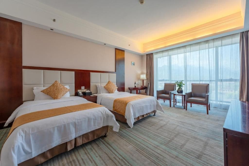 Habitación de hotel con dos camas, una zona de estar con mesa y sillas, y un ventanal con cortinas.