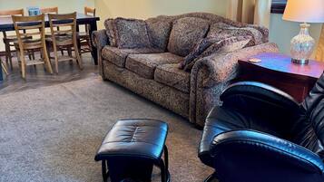 Een woonkamer met een gestreept sofa, een zwarte leren poef en een zwarte lederen fauteuil. In de achtergrond staat een eettafel met stoelen en een kroonluchter.