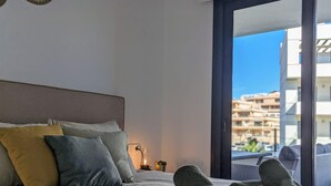 Une chambre à coucher avec un lit, des oreillers, une table de chevet, une lampe et une vue sur un immeuble par une grande fenêtre.