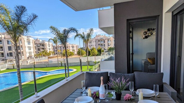 Un balcon avec une table à manger pour deux personnes, donnant sur une piscine et un espace paysager agrémenté de palmiers.