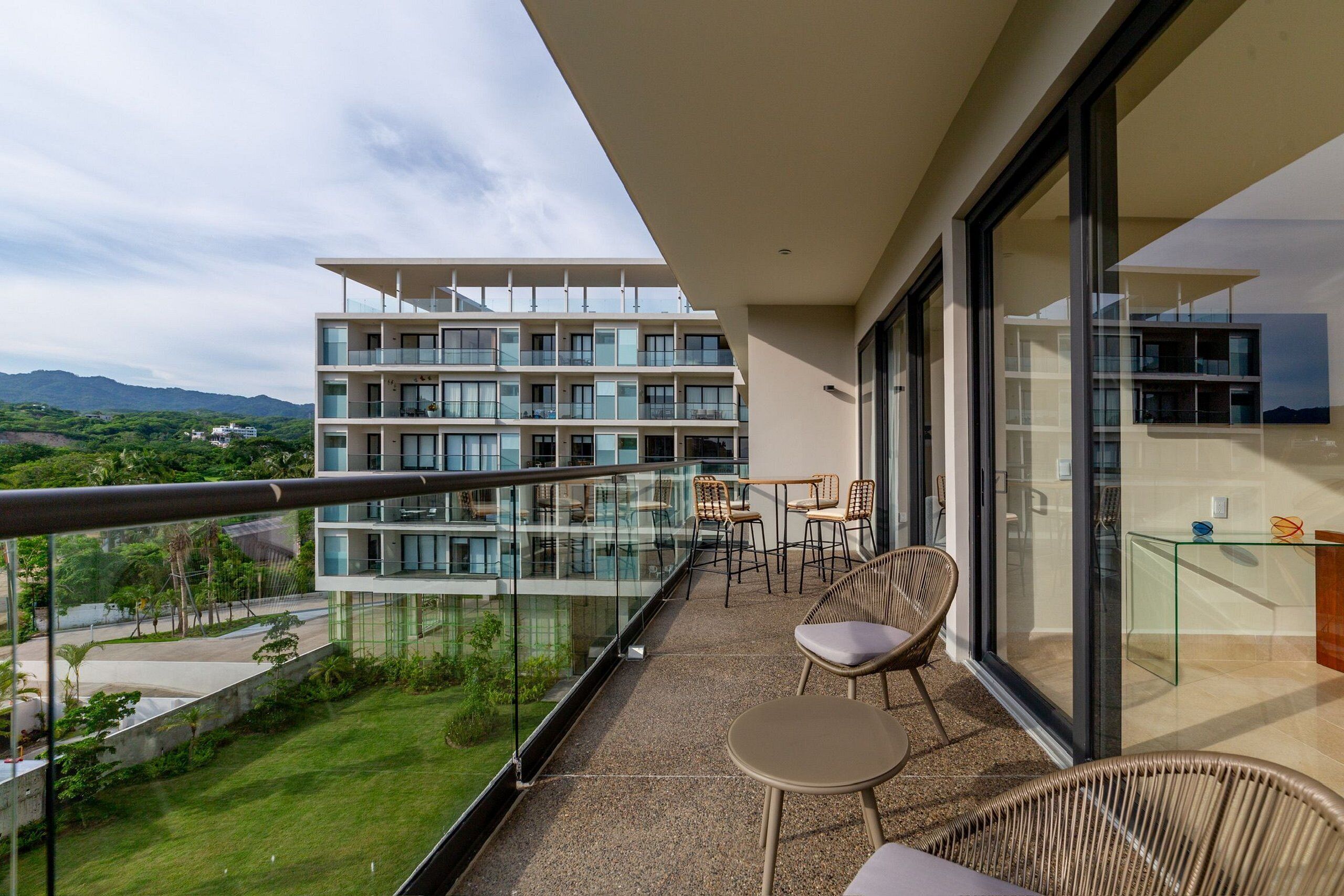 Een balkon met een glazen hekwerk, rieten meubilair en uitzicht op een meerverdiepingsgebouw en groen.