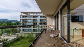 Een balkon met een glazen hekwerk, rieten meubilair en uitzicht op een meerverdiepingsgebouw en groen.