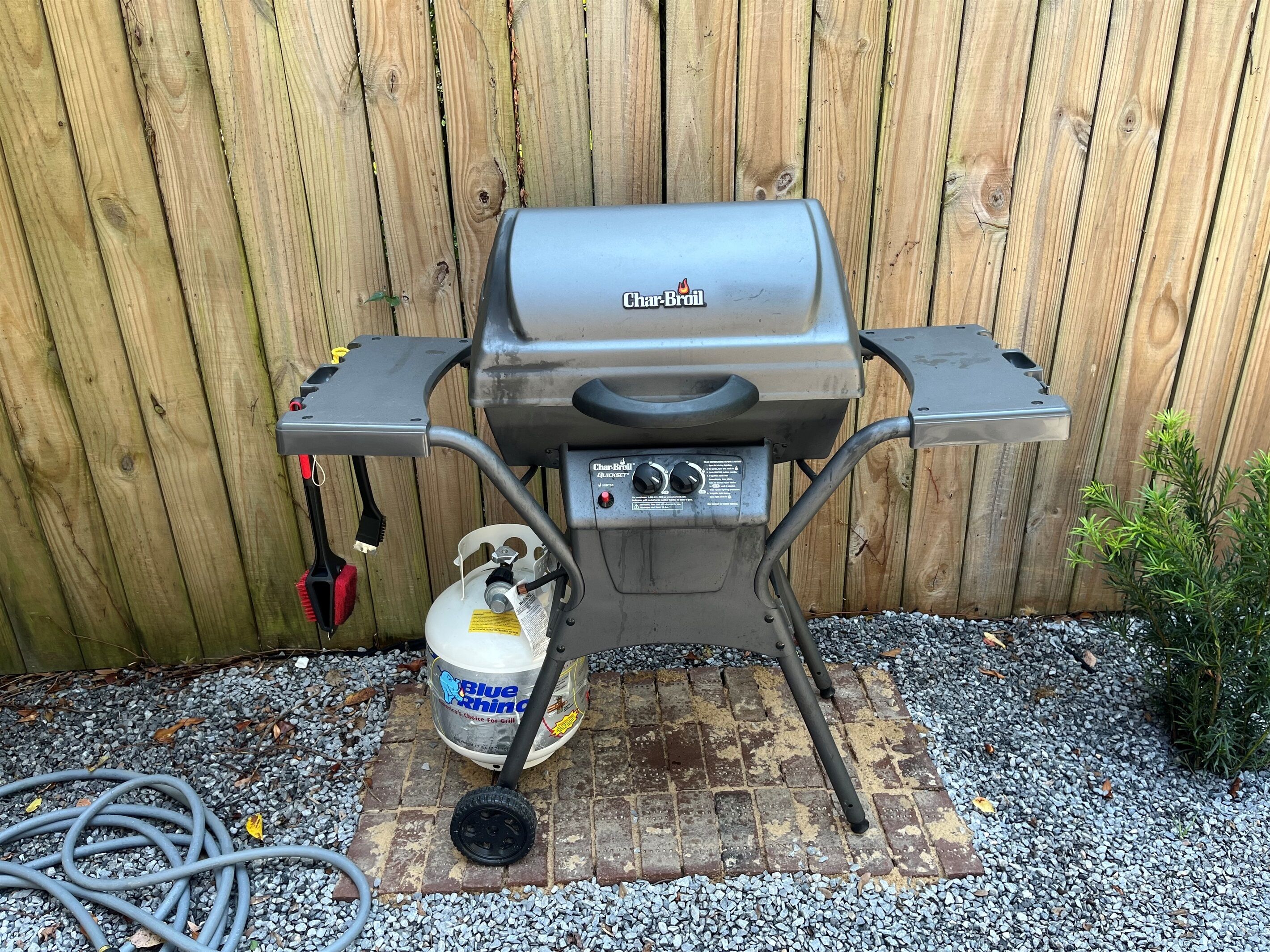 A Char-Broil gas grill with a propane tank, set on a brick base with gravel around it, against a wooden fence.