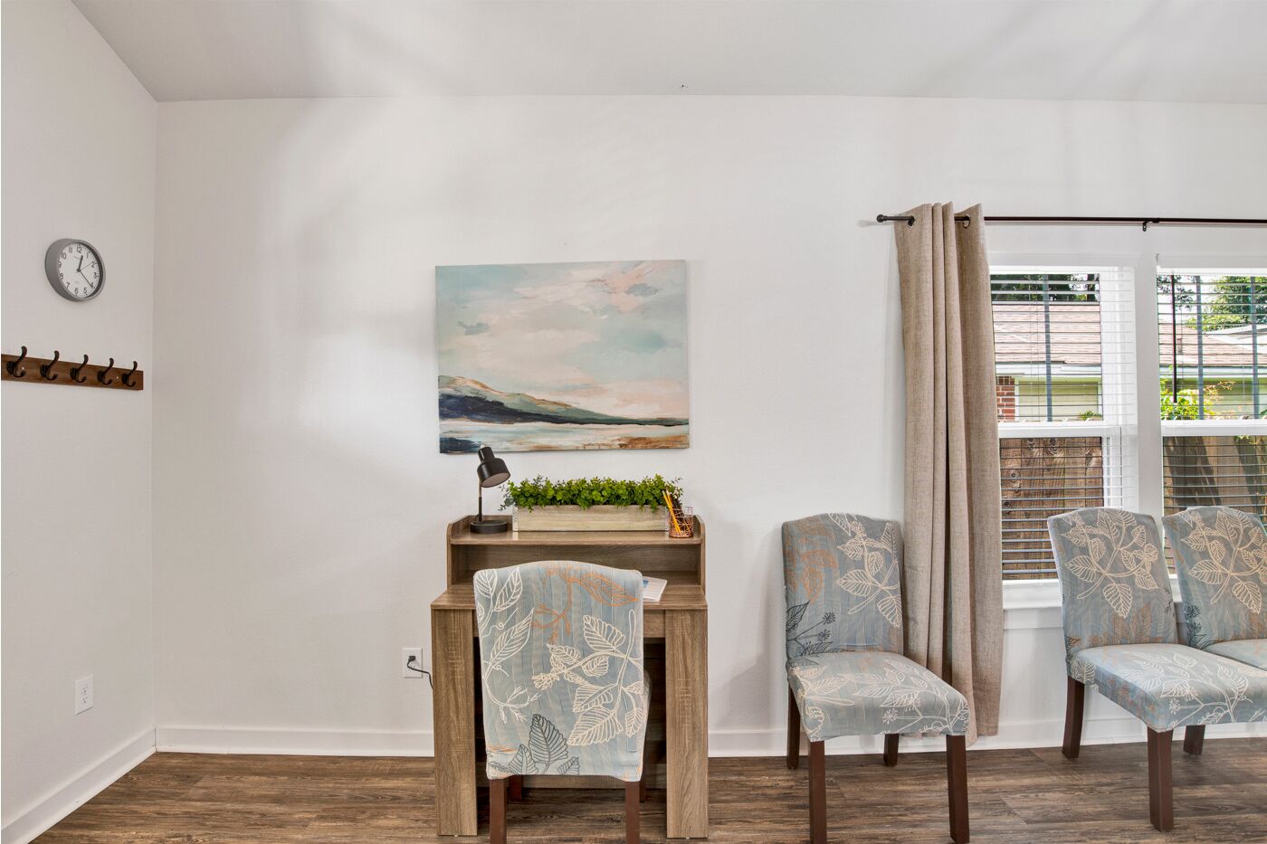 A dining area with a wooden table, two chairs with floral upholstery, a wall-mounted clock, and a painting of a landscape.