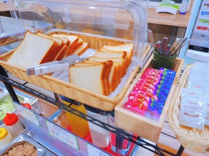 Un bufé con pan blanco en rodajas, salsas y un dispensador de bebidas de colores.