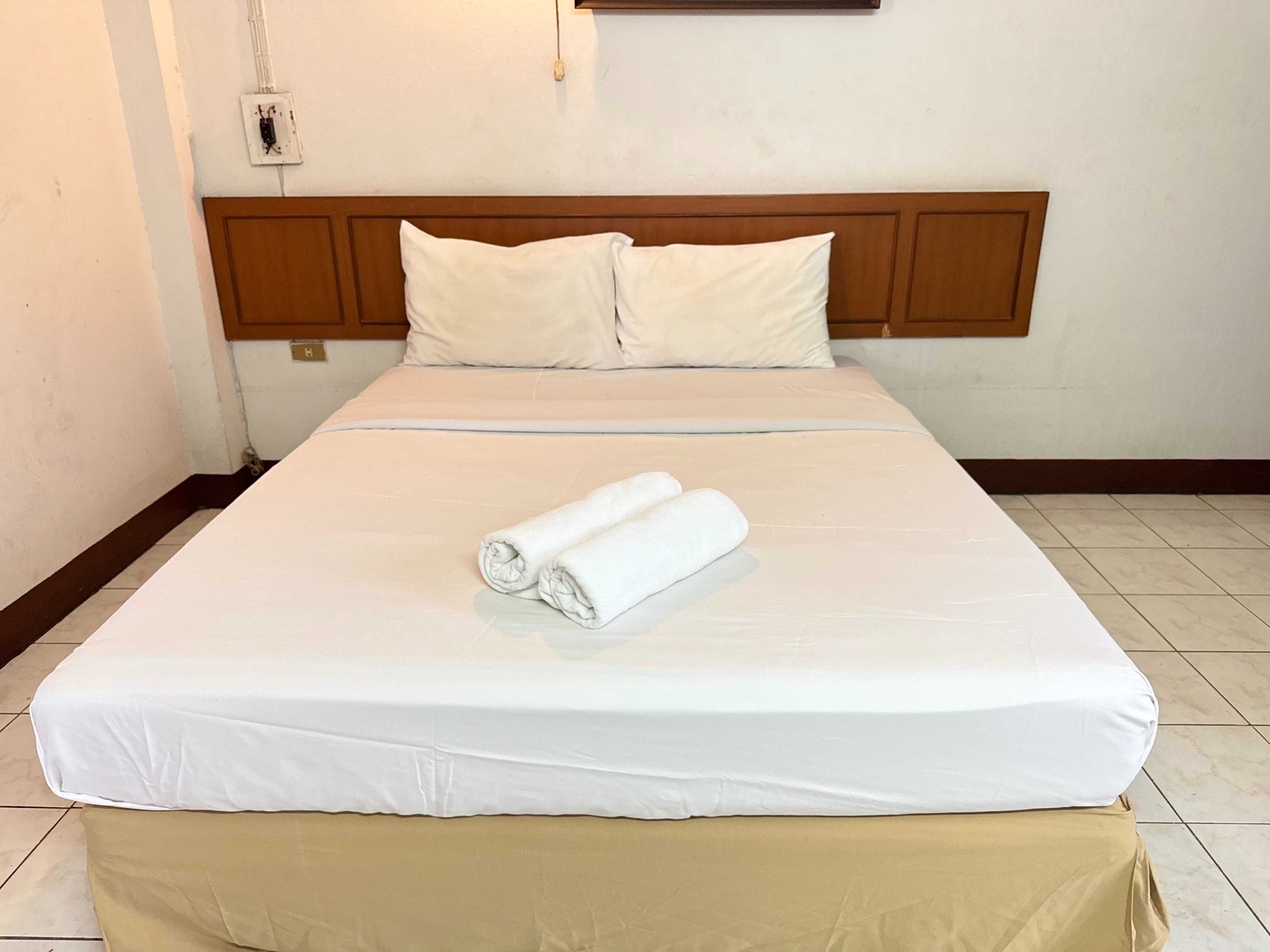 A neatly made bed with white linens and a beige bed skirt, a wooden headboard, and a white wall with a light switch.