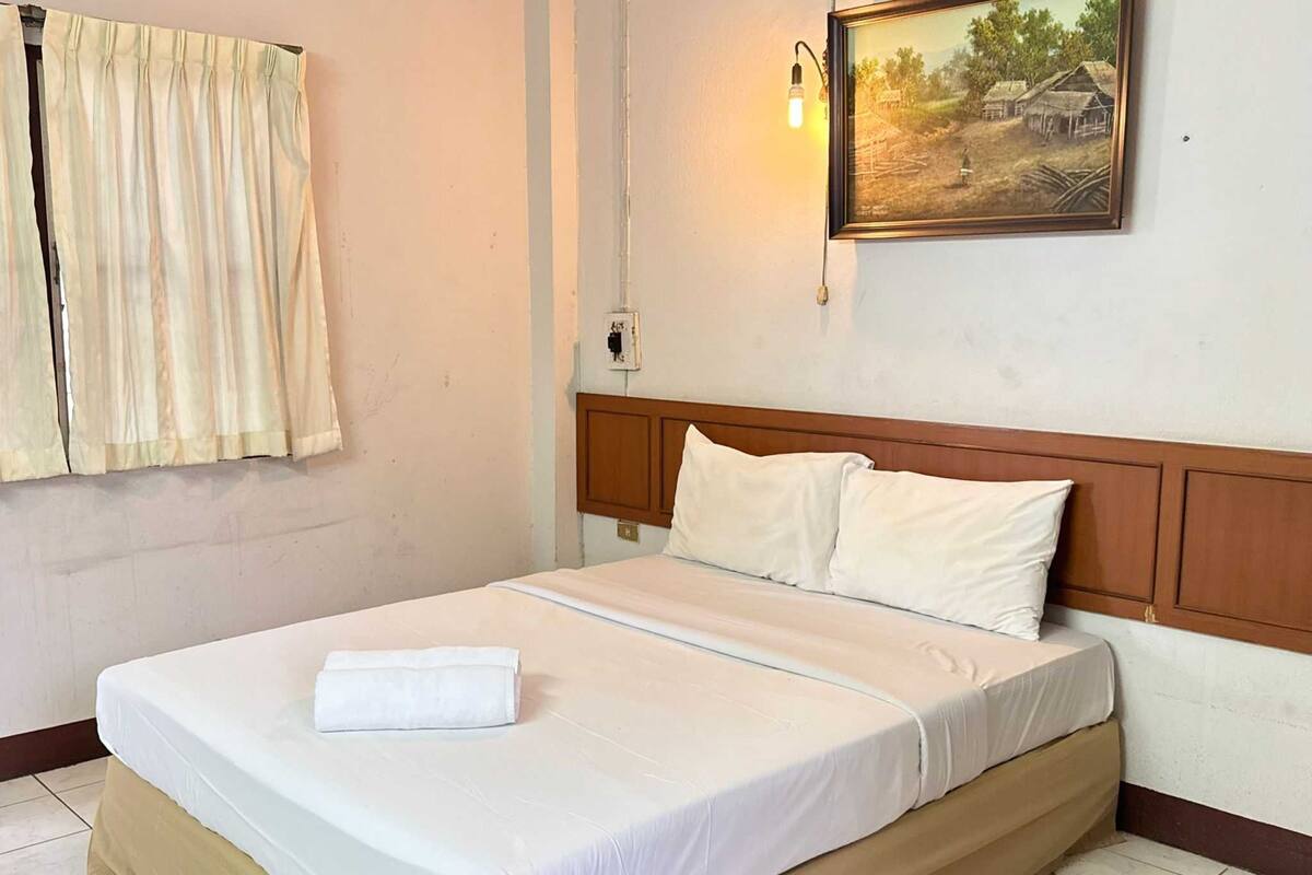 A neatly made bed with white linens and a folded towel, a wooden headboard, a wall-mounted light, and a framed picture of a rural scene.