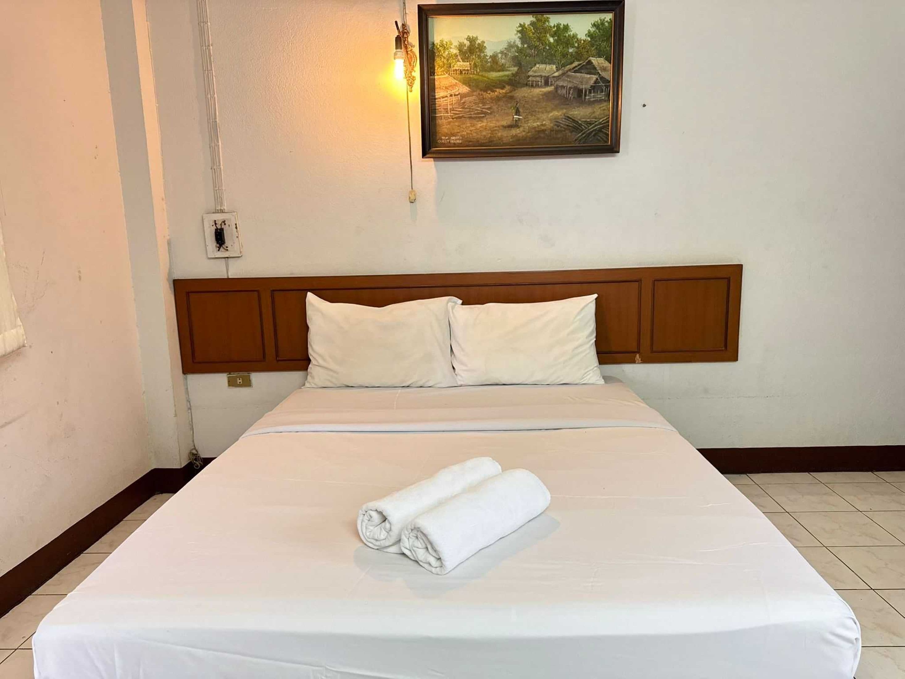 A neatly made bed with white linens and pillows, a rolled towel on the bed, and a framed painting above the headboard.