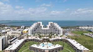 A large hotel with a central courtyard and a pool, surrounded by a coastal view and a bridge in the background.