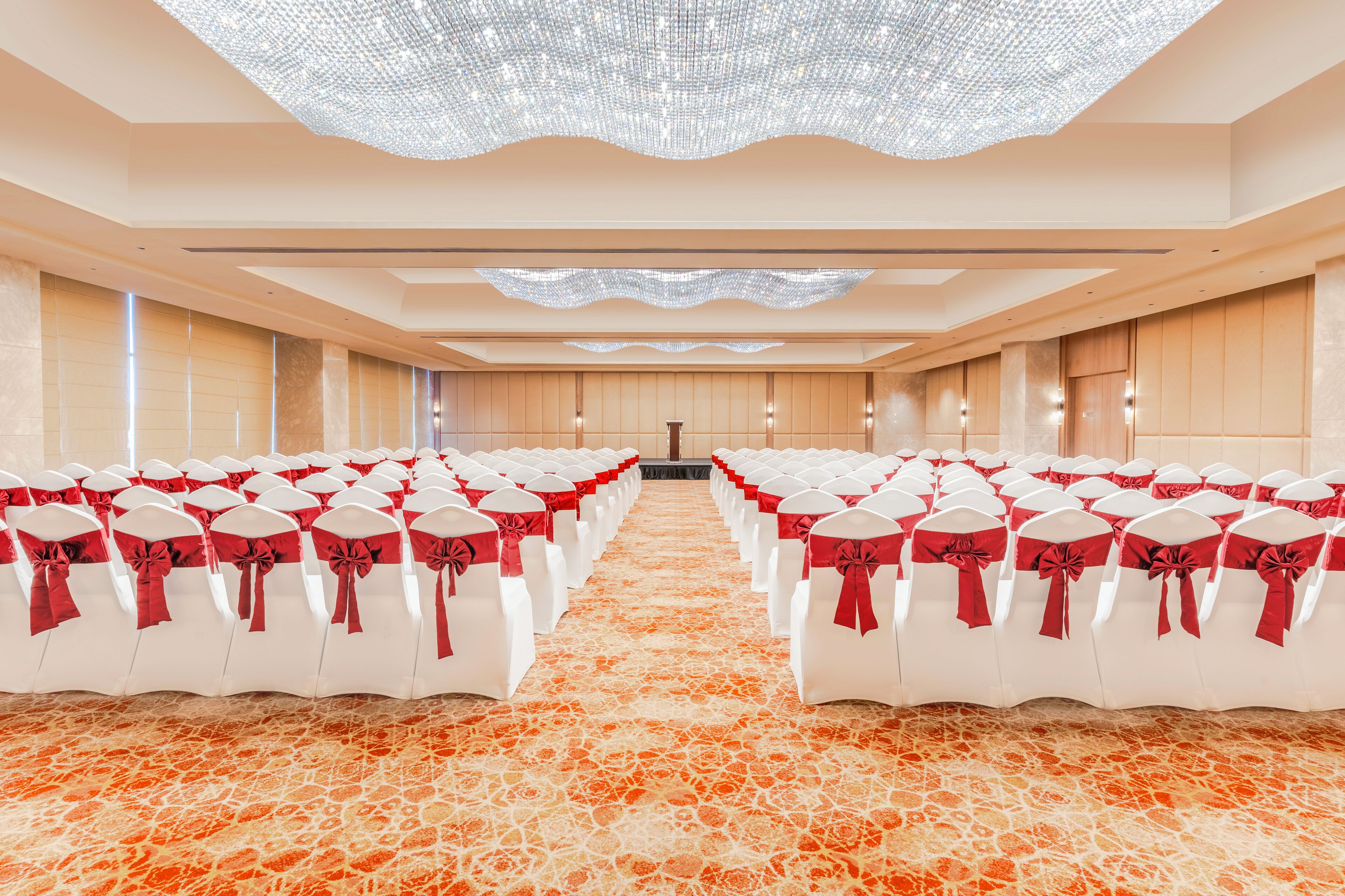 Un amplio salón con filas de sillas blancas decoradas con cintas rojas, una alfombra estampada y una gran lámpara de araña.