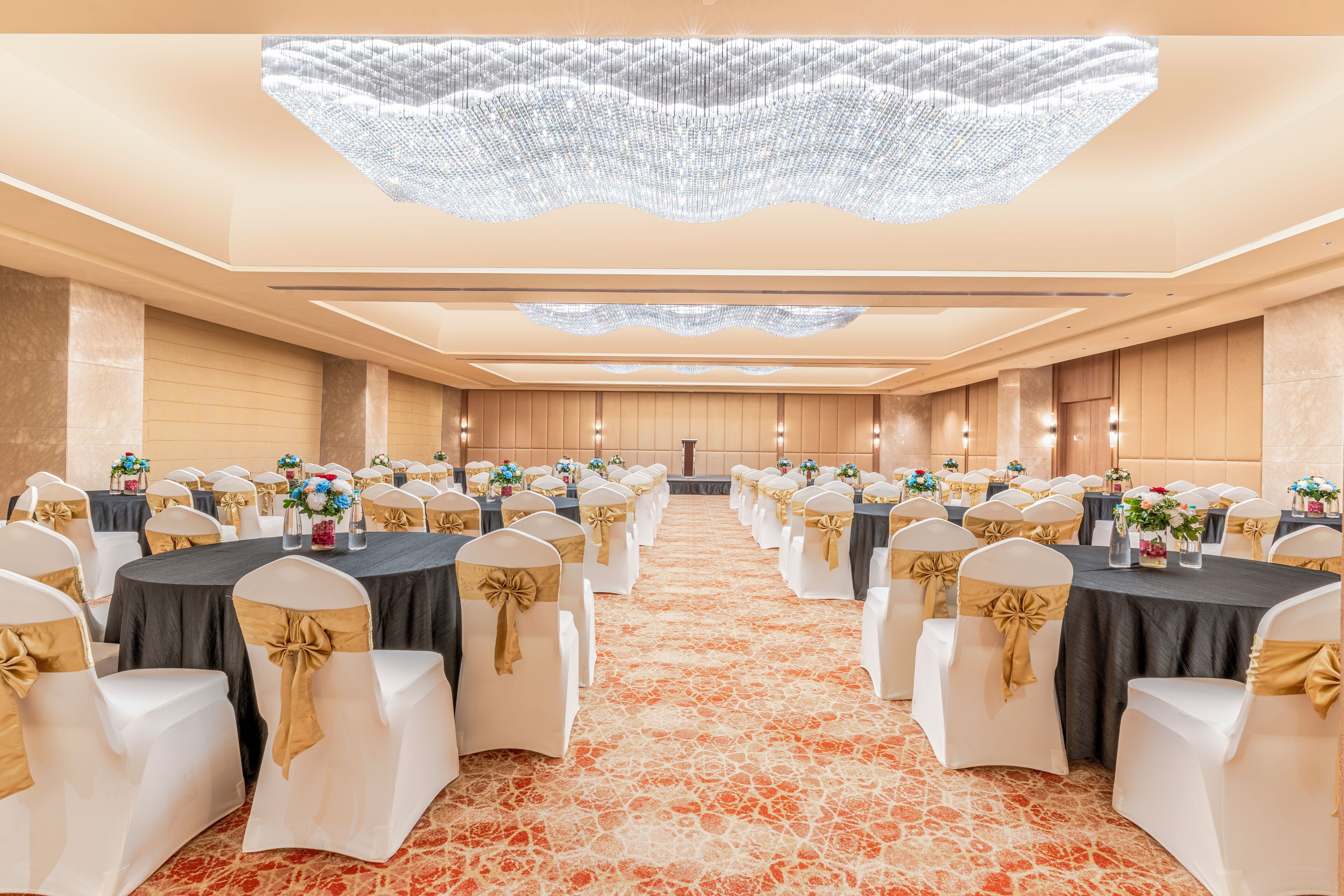 A banquet hall with round tables, white chairs with gold sashes, and a patterned carpet.