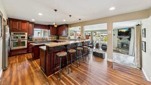 A spacious kitchen with wooden cabinets, a central island with stools, and a stone fireplace in the adjacent living area.
