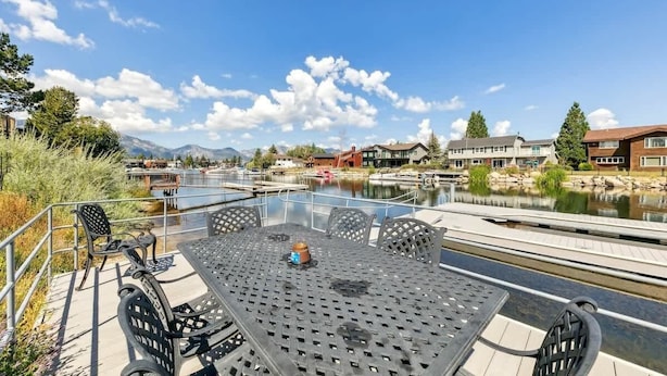 A glass table with chairs on a deck overlooking a body of water, with buildings and mountains in the background.