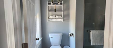 Ein Badezimmer mit Toilette, einem kleinen Mülleimer, einem Regal mit Pflanzen und einem Handtuchhalter.
