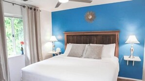 A bedroom with a large bed, two bedside tables with lamps, a ceiling fan, and a window with curtains.