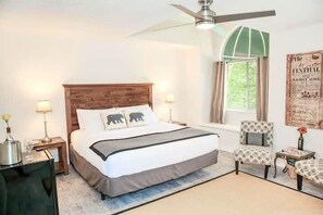 Ein Schlafzimmer mit einem hölzernen Kopfteil, zwei Nachttischlampen, einem Bett mit weißer Bettdecke und grauer Tagesdecke, einem kleinen Tisch mit einer Vase und einem Stuhl mit gemustertem Kissen.