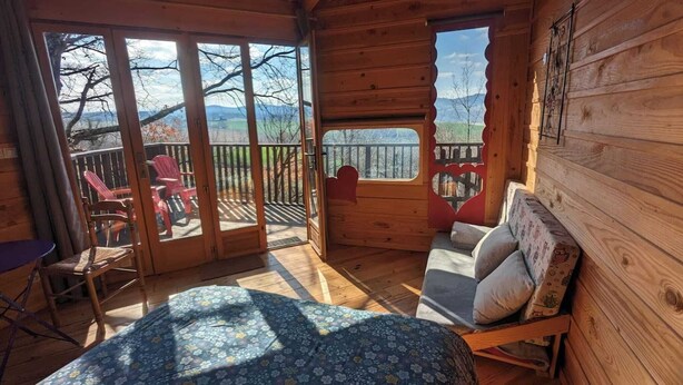 A cozy cabin room with a bed, a small table, and a chair. It features a large sliding glass door leading to an outdoor deck with chairs. - Cabanes Sainte Camelle (Saint Victor Rouzaud)
