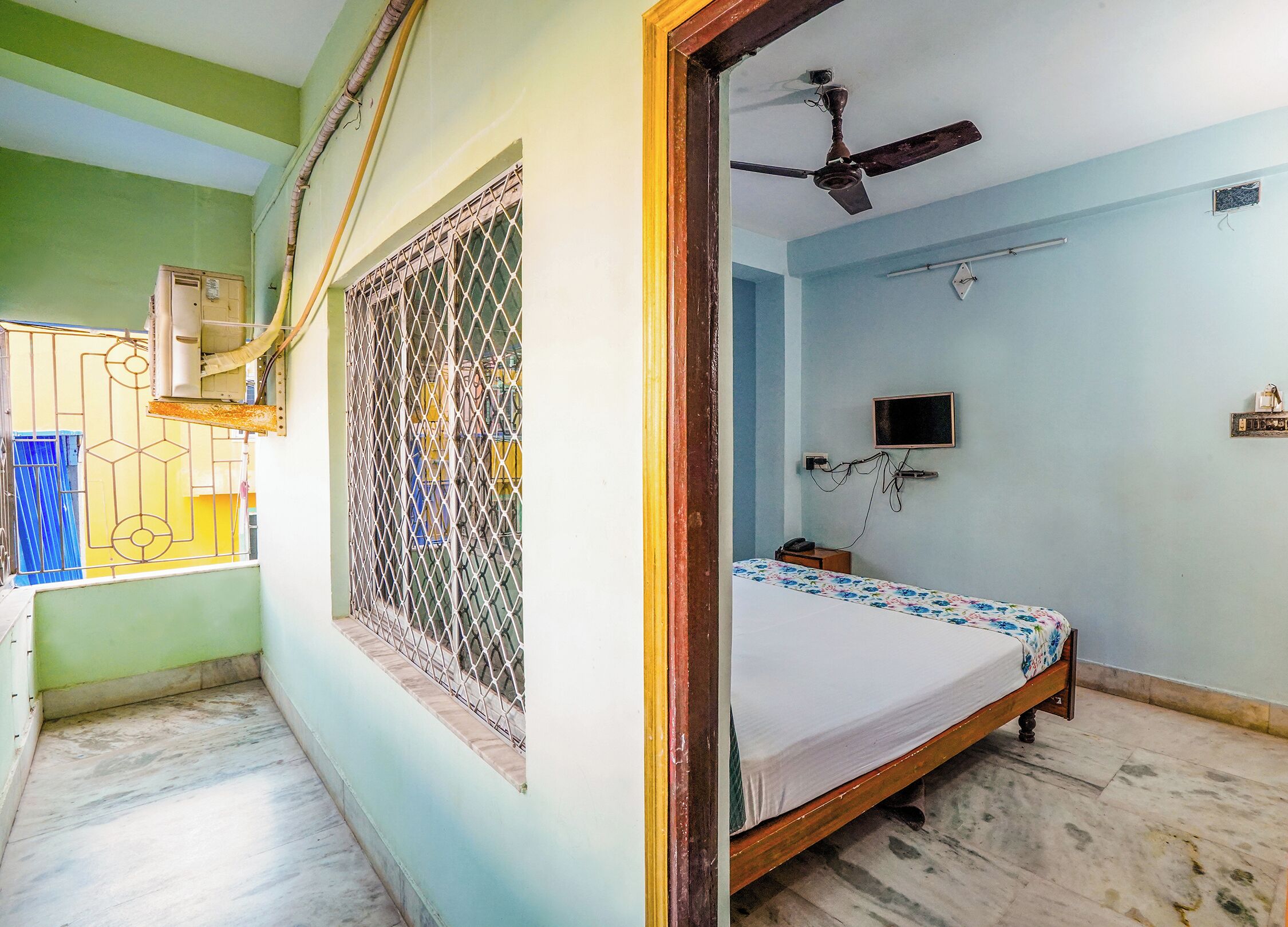 A bedroom with a bed, a ceiling fan, a TV mounted on the wall, and a window with a metal grill.