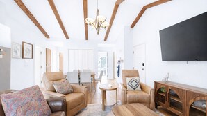 A spacious living room with high ceilings, wooden beams, a chandelier, leather sofas, a coffee table, and a flat-screen TV.