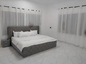 A modern bedroom with a grey tufted headboard, white bedding, and sheer curtains. - Sleepeaze Hotel (Afienya)