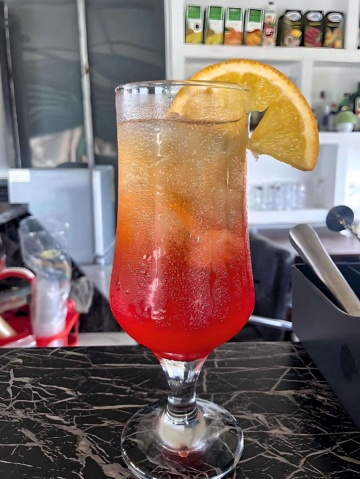 Un vaso de cóctel con un degradado de color, cubitos de hielo y una rodaja de naranja como adorno sobre una encimera de mármol.