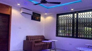 Une pièce moderne avec un canapé en cuir marron, deux petites tables rondes, un ventilateur de plafond et une grande fenêtre ornée d’une grille métallique.