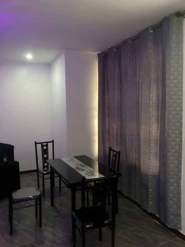 A dining area with a table and four chairs, a ceiling light, and a window with curtains. - Sleepeaze Hotel (Afienya)