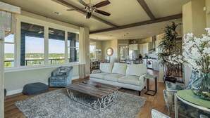 A spacious living area with a ceiling fan, large windows, a white sofa, a coffee table, and a view of the kitchen.