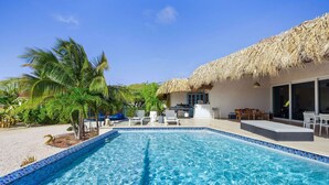 A swimming pool with a blue tiled edge, surrounded by lounge chairs and a thatched-roof building.
