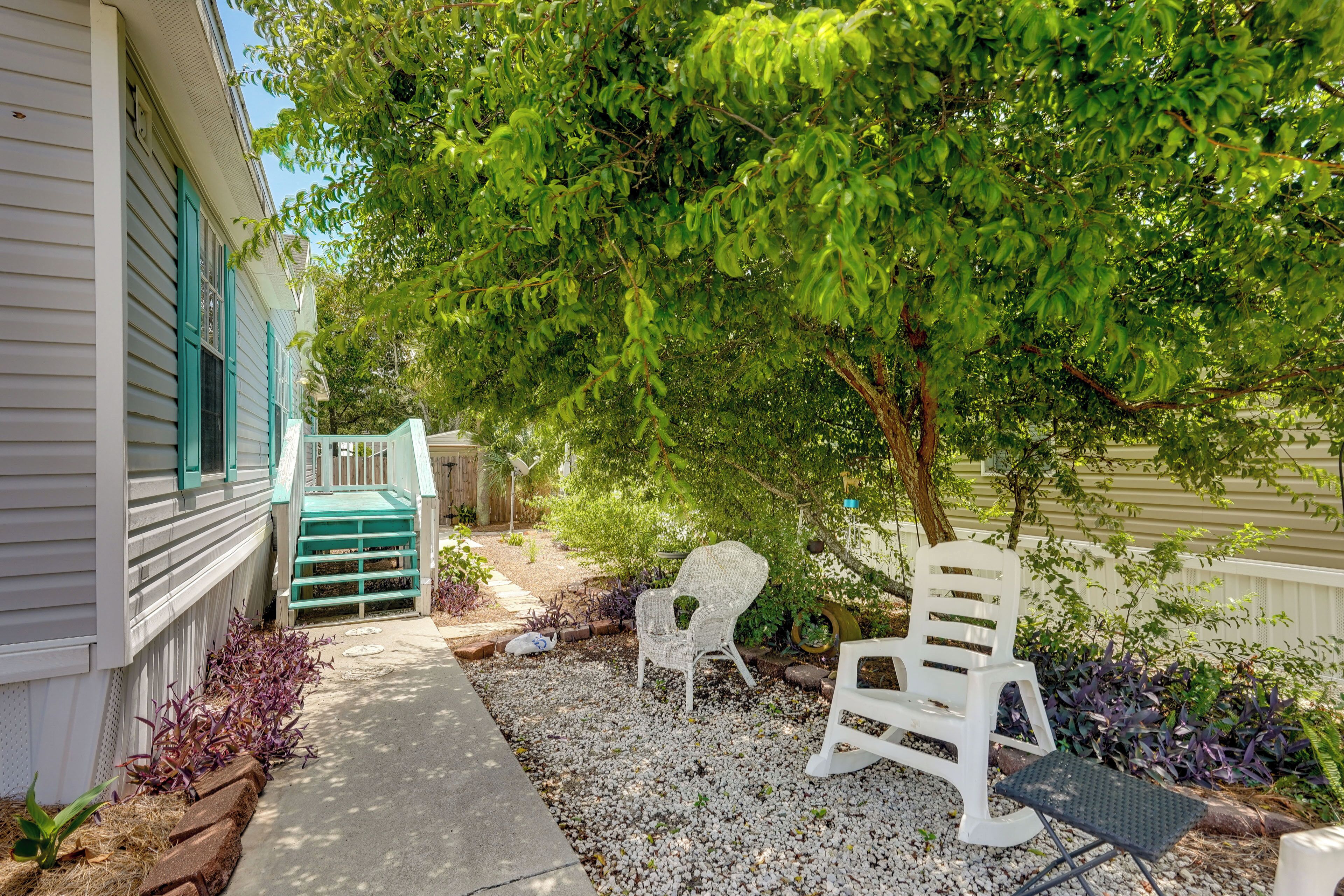 Un jardin avec un chemin de gravier, deux chaises blanches, une petite table et un arbre qui offre de l’ombre.
