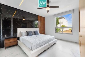 Un dormitorio moderno con una cama grande, un ventilador de techo y vista a un edificio blanco a través de una ventana.