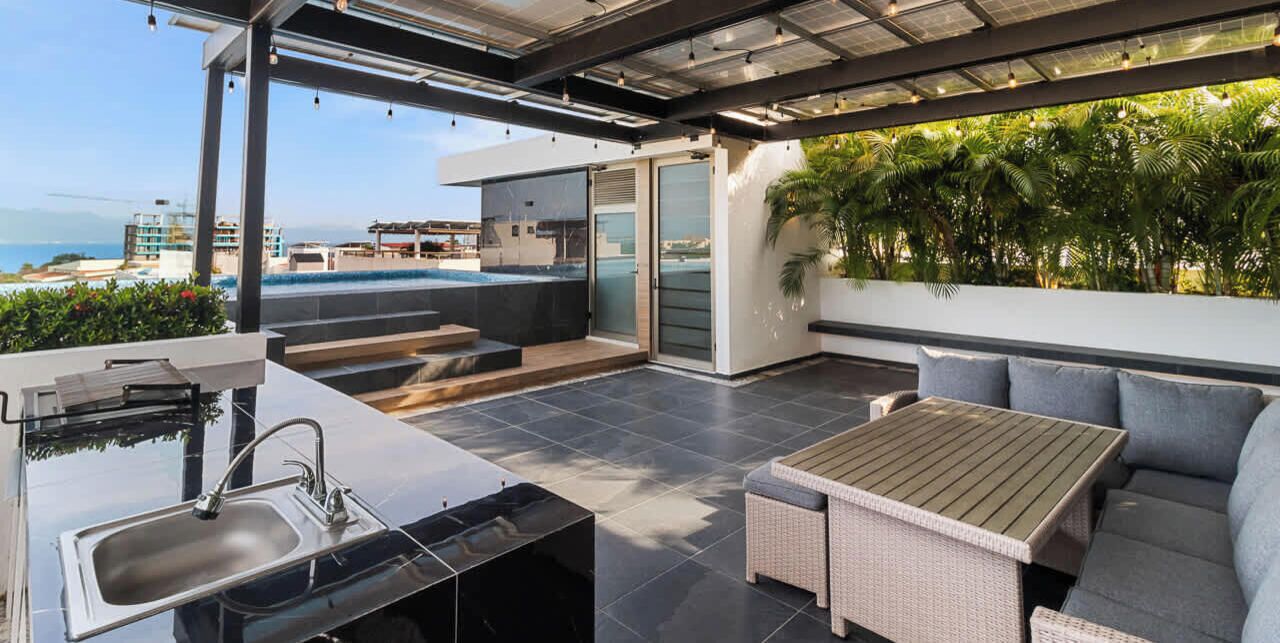 A modern outdoor living area with a kitchen, seating, and a view of the ocean.