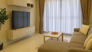 A modern living room with a flat-screen TV on a wooden stand, a beige sofa, a coffee table, and a large window with curtains.