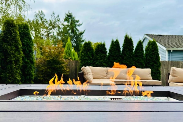 Una chimenea decorativa con canicas de vidrio en un marco metálico rectangular, un sofá exterior color beige y una valla de madera en un patio trasero residencial.