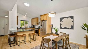 Una cocina moderna con una isla central, taburetes de madera, una mesa de comedor con sillas, un cuadro grande con motivos florales en la pared y una lámpara colgante.