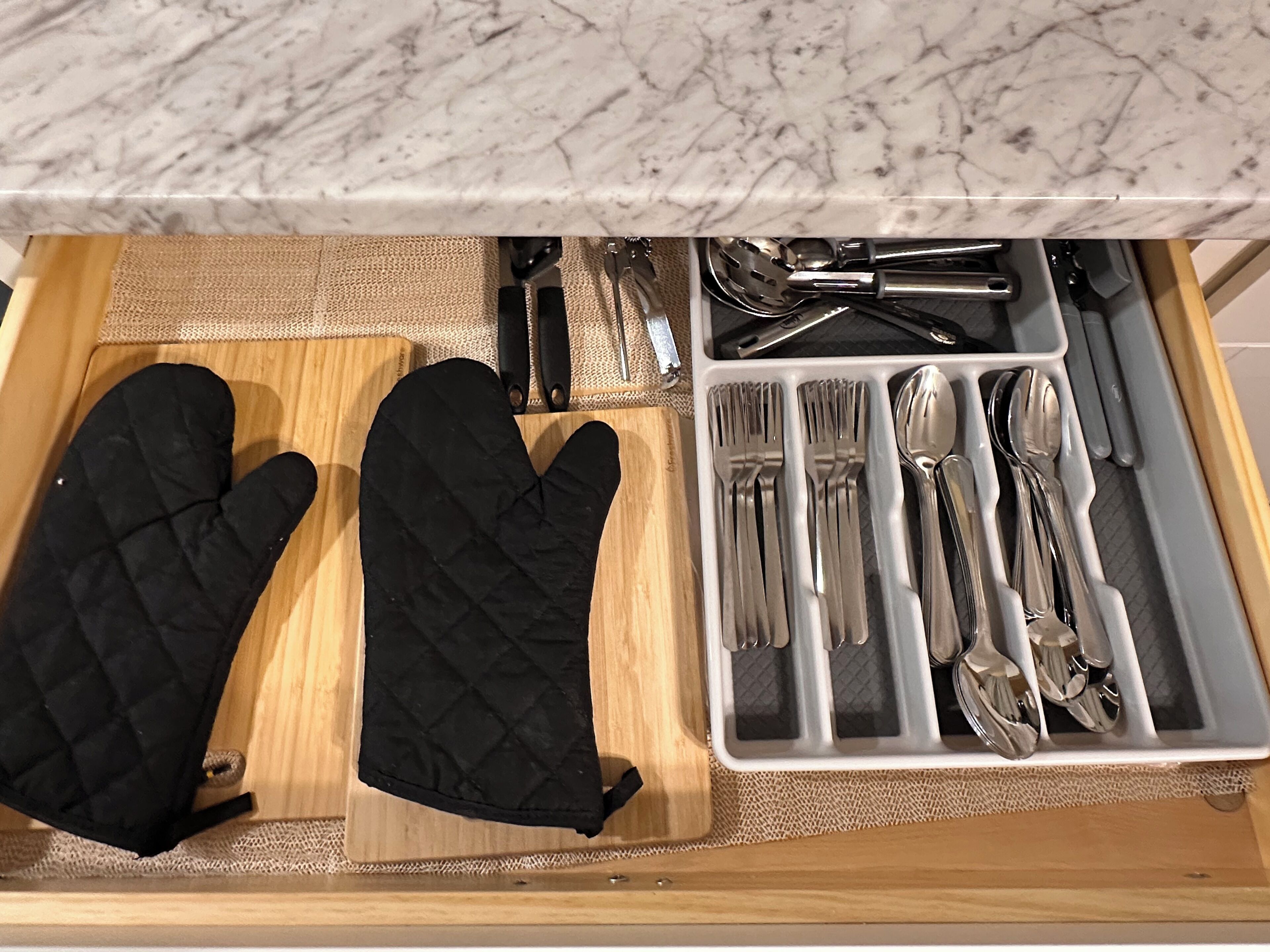 A kitchen drawer with black oven mitts, a cutting board, and a utensil organizer containing forks, knives, and spoons.