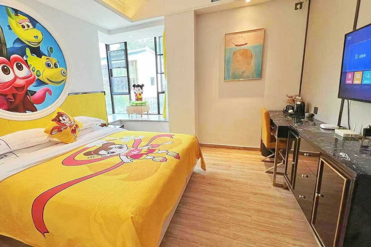 A hotel room with a bed featuring a cartoon character design, a desk with a computer, and a large window offering a view of the outside.