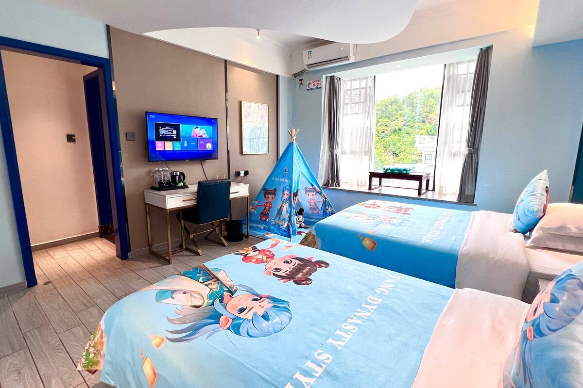 A hotel room with two beds, a desk, a TV, and a window with a view of greenery.