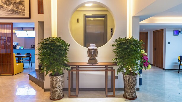 A reception area with a wooden reception desk, two potted plants, a Buddha statue, and an elevator. - Zhuhai Yujia Boutique Homestay (Zhuhai)