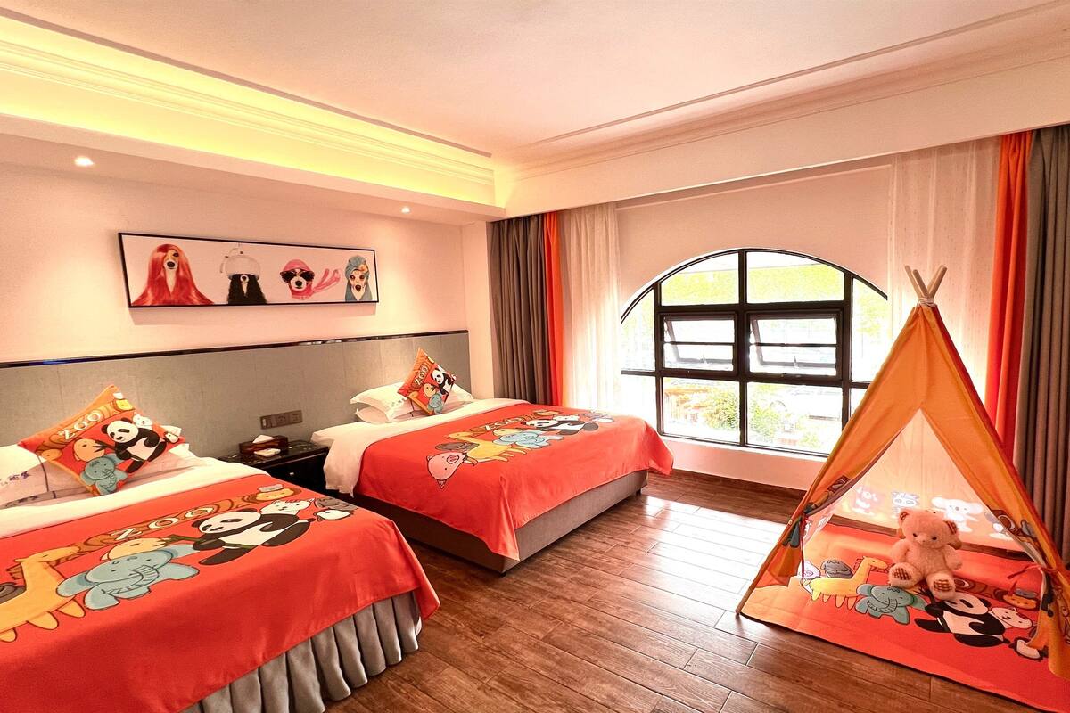 A hotel room with two beds, a window with curtains, a wall-mounted picture, and a small tent-like structure with toys.