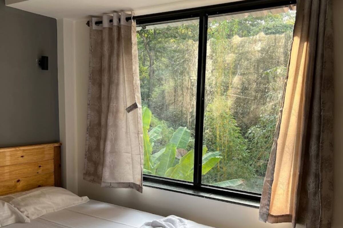 Une chambre à coucher avec un lit, une tête de lit en bois, une fenêtre avec des rideaux et une vue sur la verdure à l’extérieur.