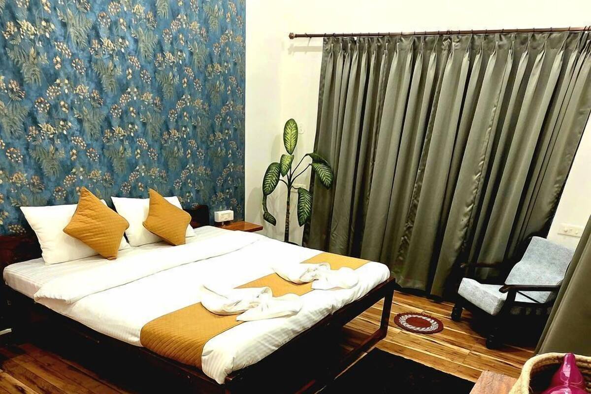 Une chambre avec un plancher en bois, un lit avec une literie blanche et deux oreillers marron, un papier peint à motifs floraux bleus, une plante en pot, une chaise grise et une table en bois avec un panier.