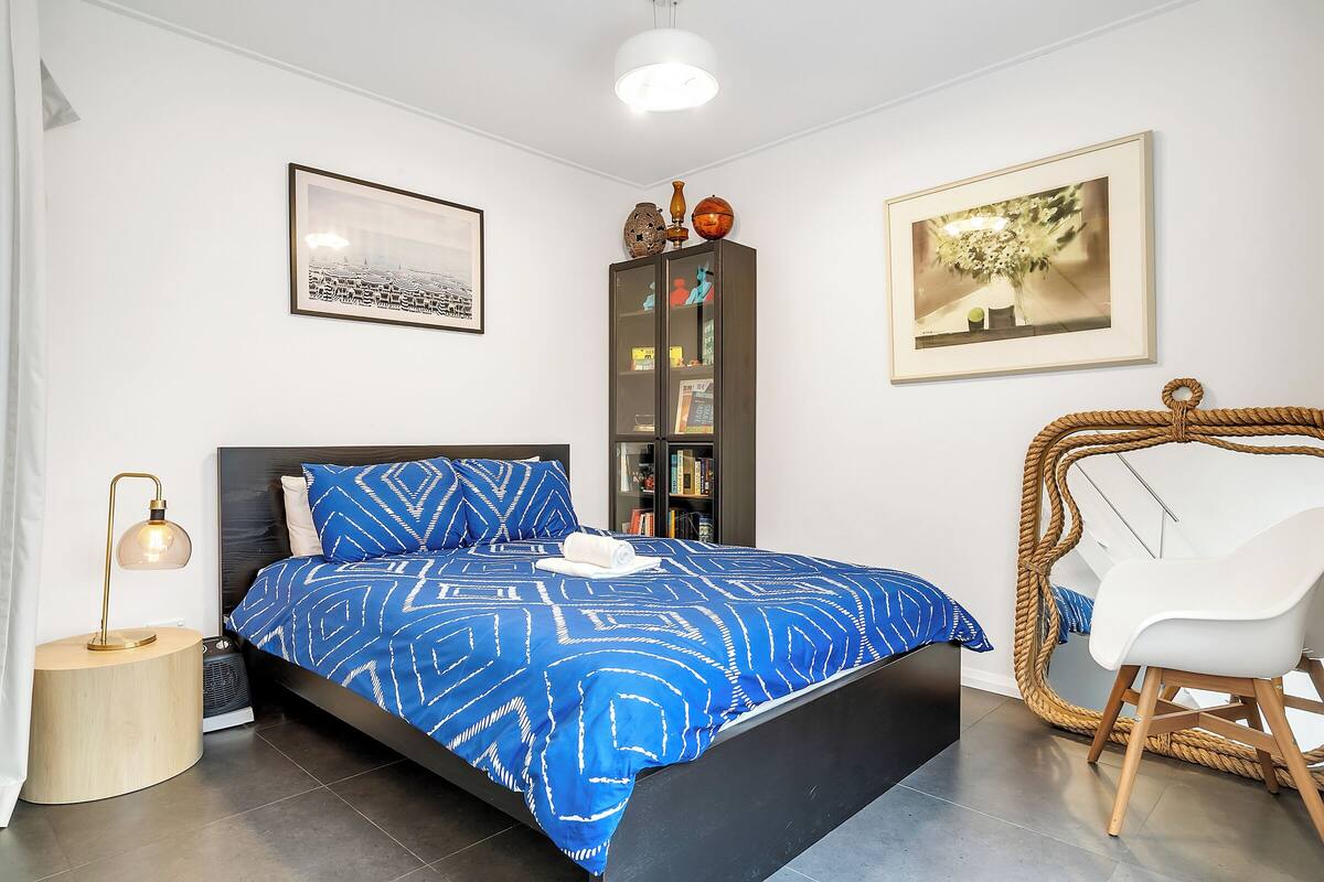 Une chambre à coucher comprenant un lit, une table de chevet, une lampe, une bibliothèque, une chaise et des œuvres d’art accrochées aux murs.