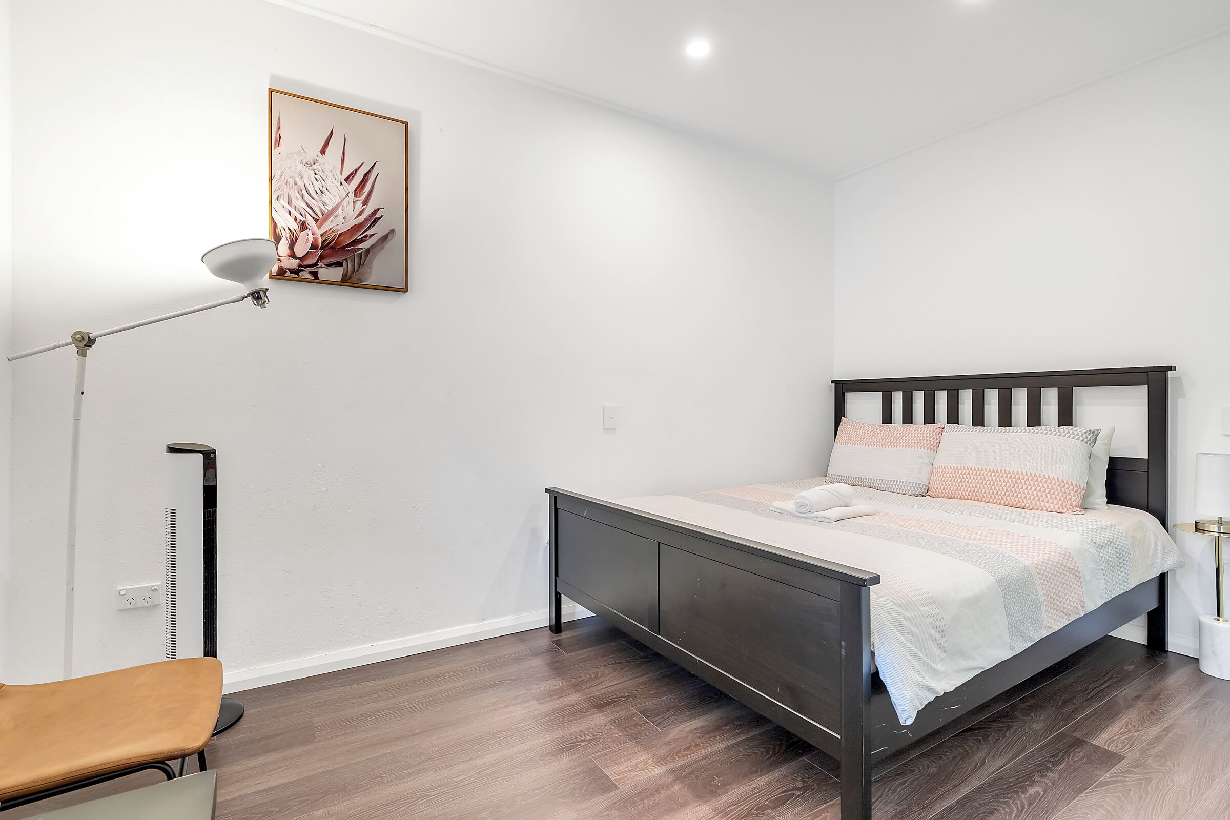 A modern bedroom with a dark wooden bed, white bedding, a floor lamp, and a wall-mounted artwork.