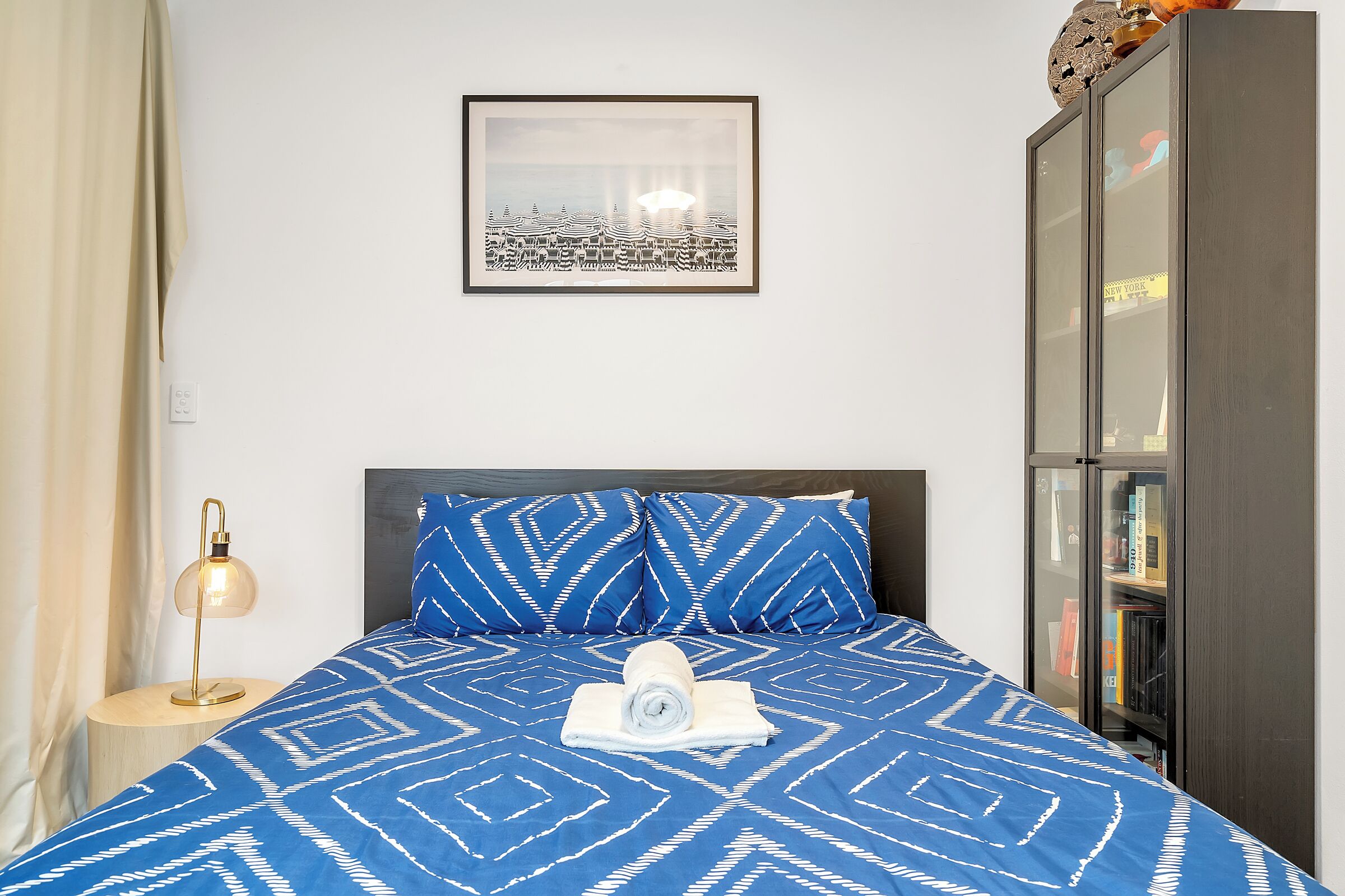 Un letto rifatto con cura, con una coperta a righe blu e bianche, un asciugamano arrotolato e cuscini coordinati. Sopra il letto è appeso un quadro incorniciato, e a destra si nota una libreria.