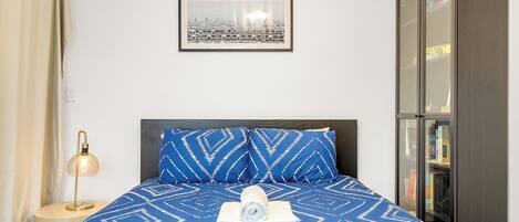 Un letto rifatto con cura, con una coperta a righe blu e bianche, un asciugamano arrotolato e cuscini coordinati. Sopra il letto è appeso un quadro incorniciato, e a destra si nota una libreria.