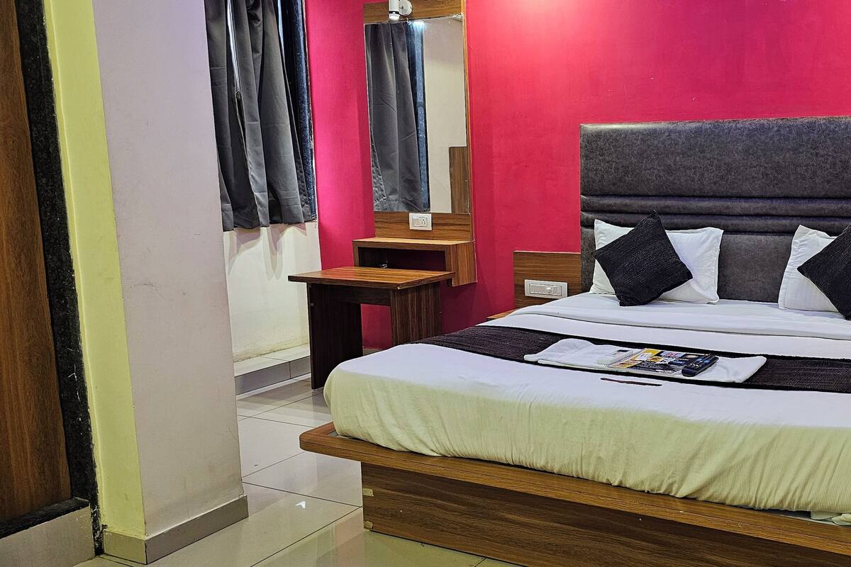 A neatly made bed with a grey headboard, white bedding, and black pillows. There is a bedside table with a lamp and a phone. The room has a red wall and a mirror above the bed.